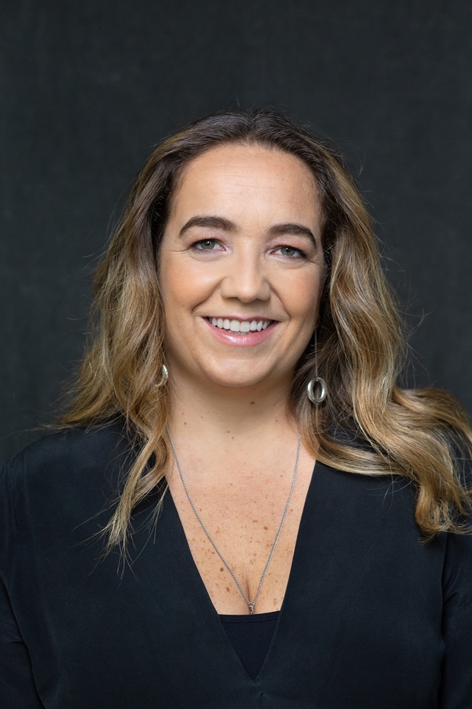 Marcia Esteves mulher branca de cabelos castanho-claros aparece em foto de close usando blusa preta com decote V e brincos de argola