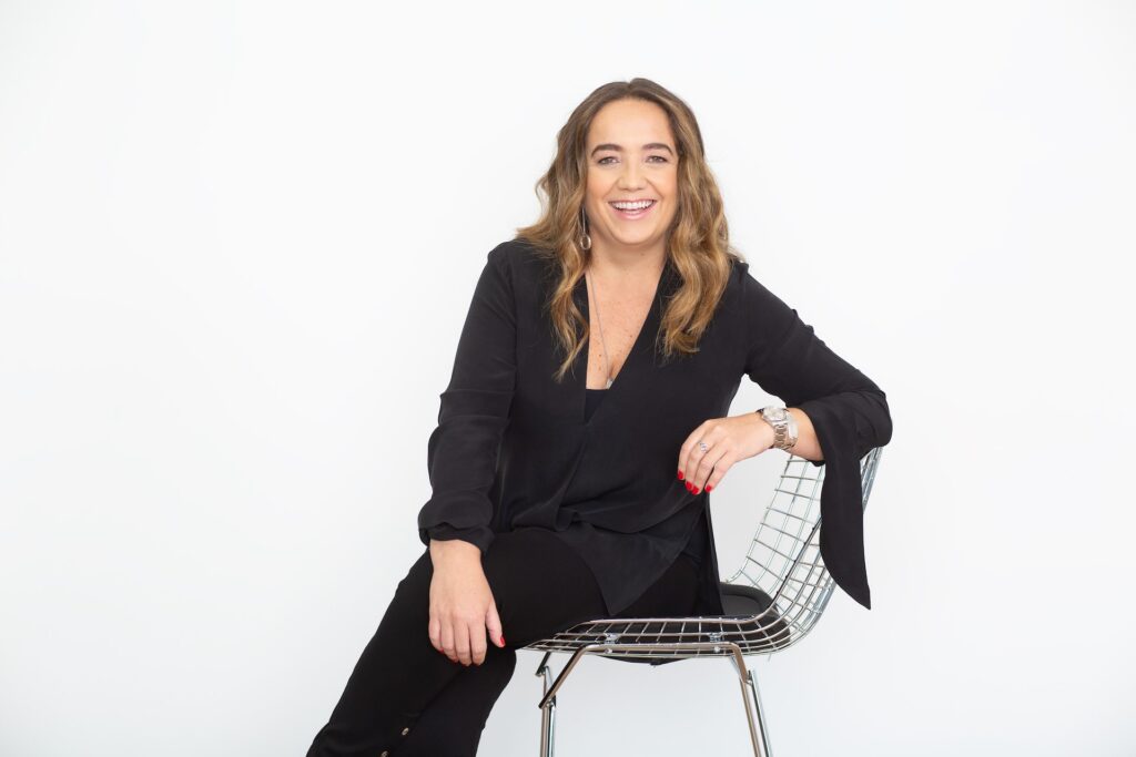 Marcia Esteves presidente da ABAP mulher branca de cabelos onsulados e longos, cstanhos, veste calça e blusa de mangas compridas preta e está sentada em cadeira de metal prateado em fundo branco